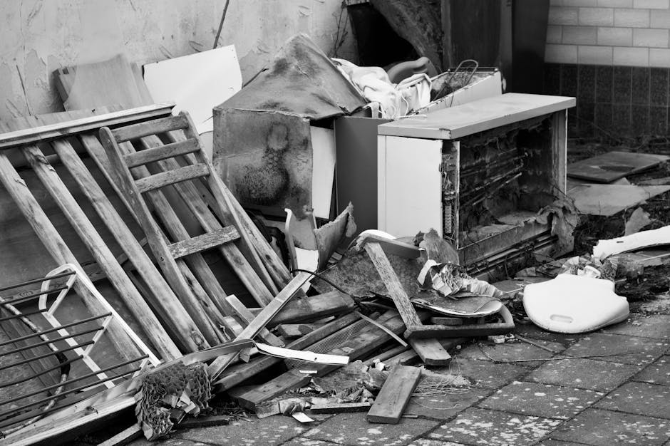 Black and white image depicting a cluttered outdoor area with discarded household items including wooden pallets, broken furniture, a damaged microwave, and various debris scattered on a paved surface