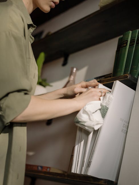 A woman wearing light green cleaning gloves and a beige shirt is dusting a wooden shelf in a room with a brick wall background. She is wiping the surface of a stack of old hardcover books, ensuring th