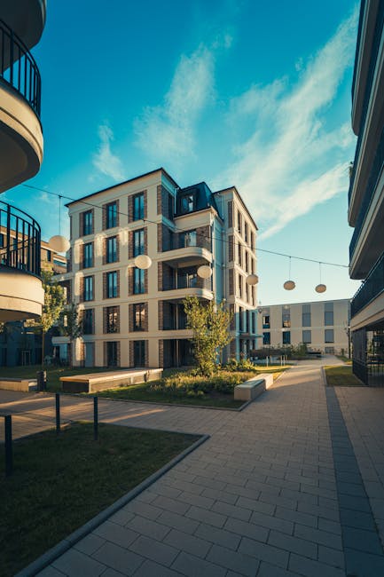 A clear outdoor communal area within a residential complex featuring paved walkways, neatly maintained green lawns, and modern multi-story buildings with large glass windows and balconies. The surfaces of the paved paths and benches appear clean and free of debris, with bright lighting from natural sunlight and decorative spherical lanterns hanging across the courtyard. The image reflects a well-maintained communal space suitable for surface cleaning and sanitation by professional cleaning services like those offered by Cleaners Pimlico, as highlighted in the Pimlico Estate & Warwick Square: communal area cleaning guide.