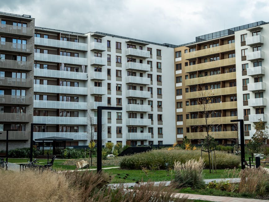 Exterior view of a modern residential apartment complex featuring three multi-storey buildings with white, beige, and grey facades, each equipped with balconies and large windows. The communal outdoor area includes well-maintained lawns, ornamental grasses, small trees, and paved walkways, with black metal arch structures and benches visible along the pathways. The scene is captured in natural daylight with overcast skies, highlighting the clean and organized appearance of the outdoor space. Cleaners Pimlico specializes in surface cleaning and maintenance of communal areas, ensuring these shared spaces remain hygienic and visually appealing, consistent with the guidelines in the Pimlico Estate & Warwick Square: communal area cleaning guide.