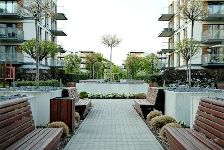 A clear outdoor communal area within a residential complex featuring paved walkways, neatly maintained green lawns, and modern multi-story buildings with large glass windows and balconies. The surfaces of the paved paths and benches appear clean and free of debris, with bright lighting from natural sunlight and decorative spherical lanterns hanging across the courtyard. The image reflects a well-maintained communal space suitable for surface cleaning and sanitation by professional cleaning services like those offered by Cleaners Pimlico, as highlighted in the Pimlico Estate & Warwick Square: communal area cleaning guide.