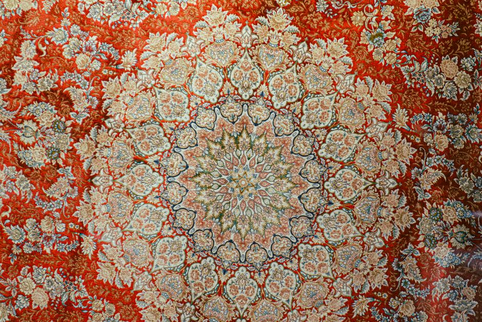 Close-up view of the corner of an ornate, traditional area rug with intricate floral patterns in shades of beige, red, and black, placed on a wooden floor with a warm, reddish-brown finish. The rug's